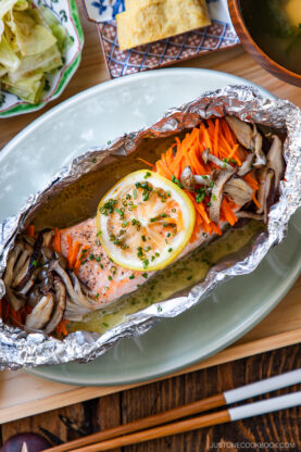 Salmon fillet baked in foil with sliced lemon, julienned carrots, and mushrooms, garnished with chives. Japanese side dishes, rice, and chopsticks are visible in the background.