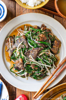 A plate of stir-fried beef liver with green vegetables and bean sprouts in a brown sauce, served on a light-colored dish with chopsticks beside it. Rice and soup bowls are partially visible in the background.