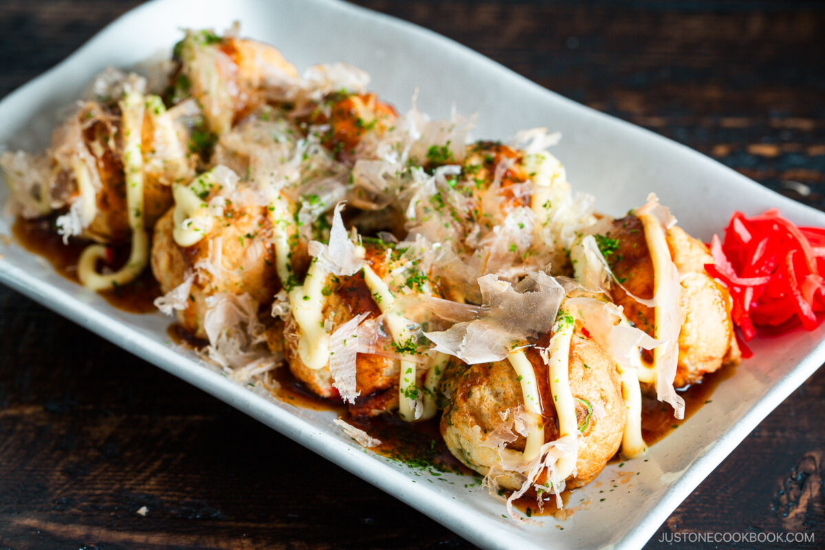 A rectangular plate of takoyaki, Japanese octopus balls, topped with mayonnaise, takoyaki sauce, bonito flakes, seaweed, and garnished with red pickled ginger on the side.