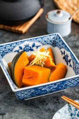 A blue and white patterned bowl filled with simmered kabocha squash pieces, topped with thinly sliced fresh ginger, sits on a dark textured surface with chopsticks and a ceramic container nearby.