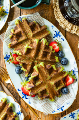 A plate with matcha waffles topped with fresh strawberries and blueberries, served on a decorative newspaper-style paper. A fork and knife are beside the plate on a wooden table.