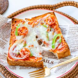 A slice of toast topped with melted cheese, cherry tomatoes, onions, and green peppers on a decorative plate, served with a gold fork on a wicker tray lined with newspaper-style parchment.