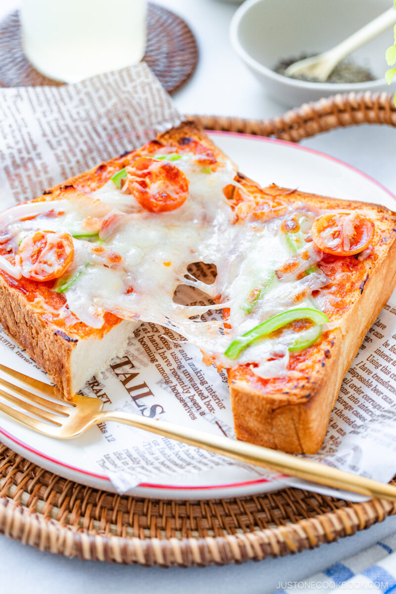 Thick toast topped with melted cheese, cherry tomatoes, and green peppers on a plate lined with newspaper-patterned paper, served with a fork and a drink in the background.
