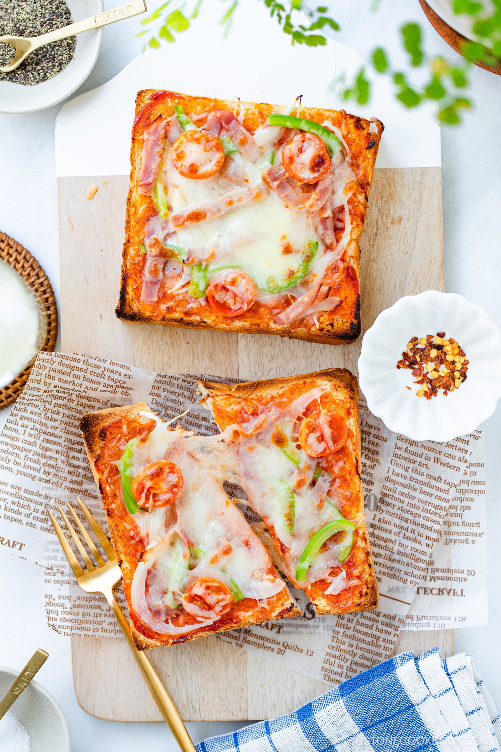 Two slices of homemade square pizza bread topped with melted cheese, cherry tomatoes, green peppers, and ham are served on a wooden board lined with newspaper-style parchment. A fork and seasonings sit nearby.
