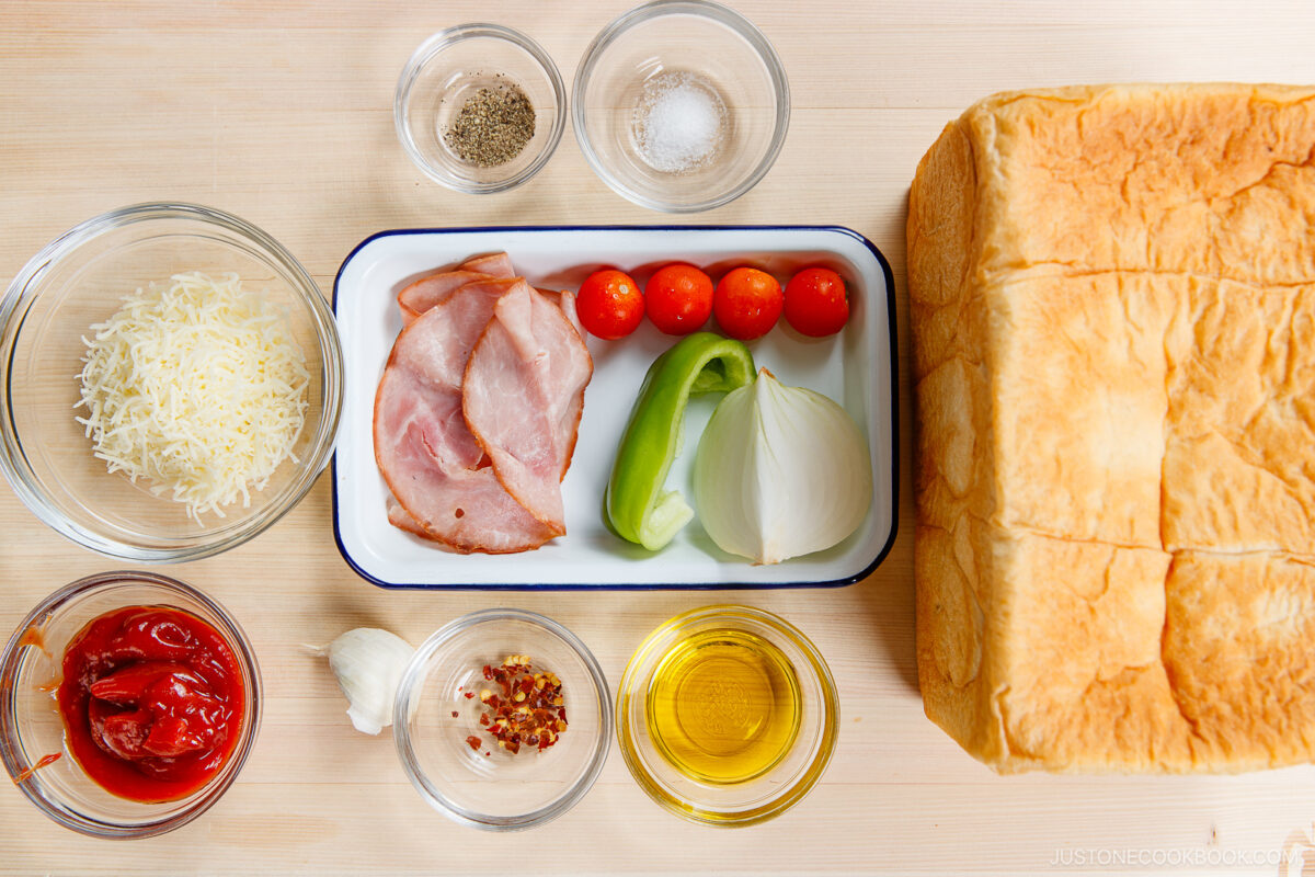 A top-down view of sandwich ingredients on a wooden surface: a loaf of bread, ham, green pepper, onion, cherry tomatoes, shredded cheese, ketchup, olive oil, a garlic clove, red pepper flakes, salt, and black pepper.