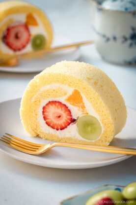 A slice of Japanese fruit roll cake filled with whipped cream, strawberry, orange, and green grape, served on a white plate with a gold fork. A teacup and another cake slice are blurred in the background.