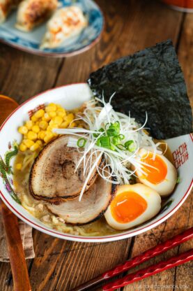 A bowl of ramen with sliced pork, soft-boiled eggs, corn, nori seaweed, green onions, and shredded white onions, served in a decorative bowl on a wooden table with red chopsticks.