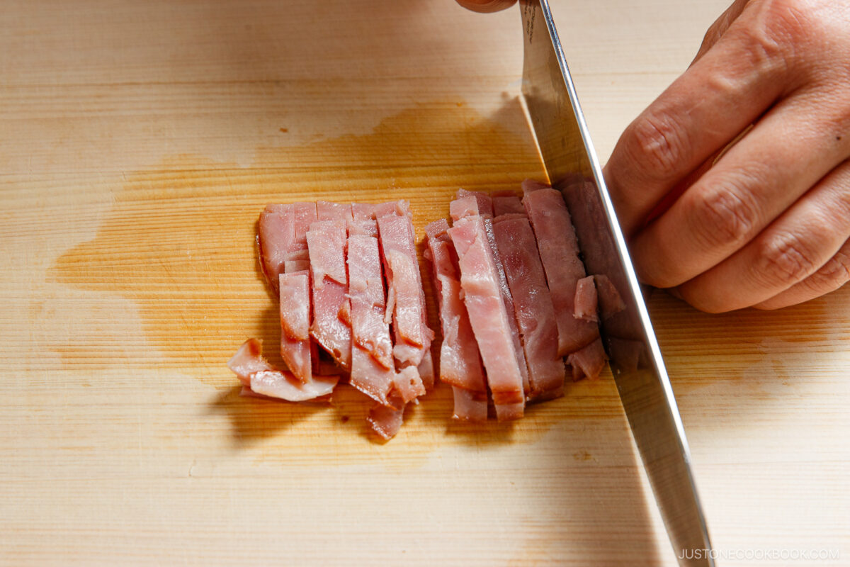 A hand holding a knife slices cooked ham into thin strips on a wooden cutting board.