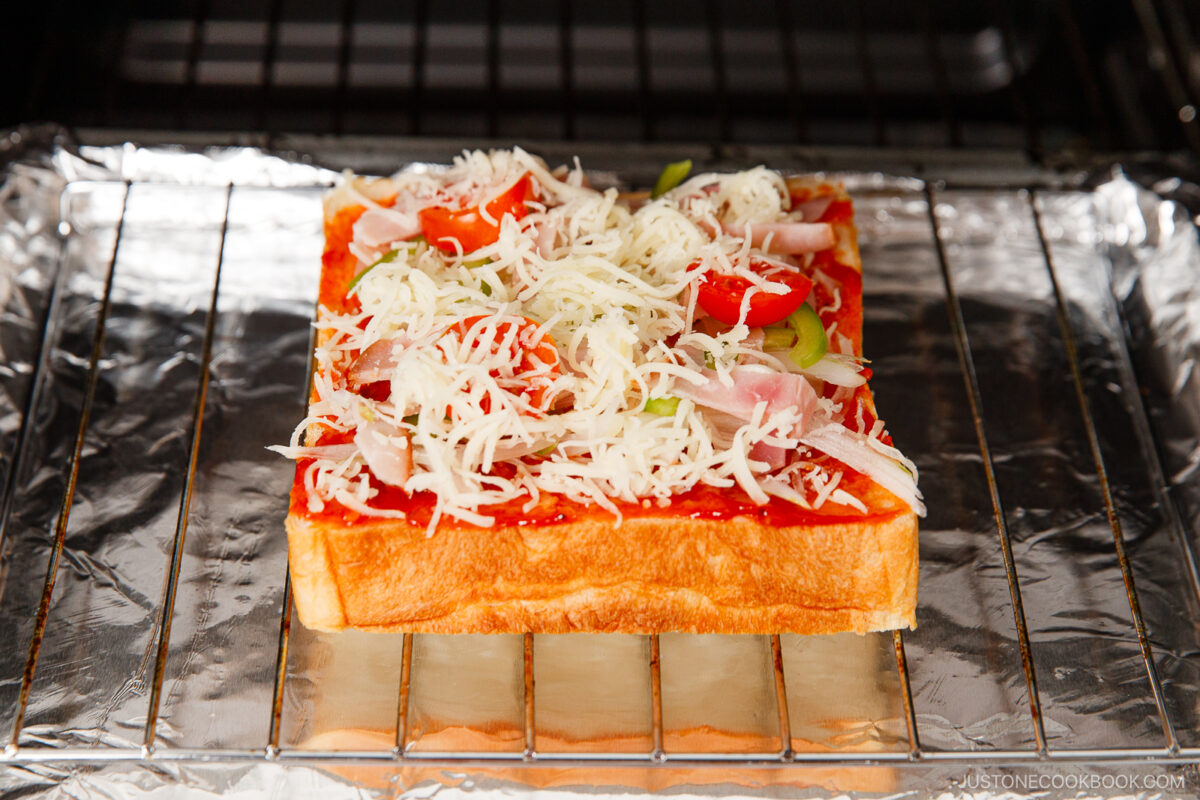A thick slice of bread topped with tomato sauce, shredded cheese, sliced ham, green peppers, and onions sits on a wire rack lined with foil inside an oven, ready to be baked.