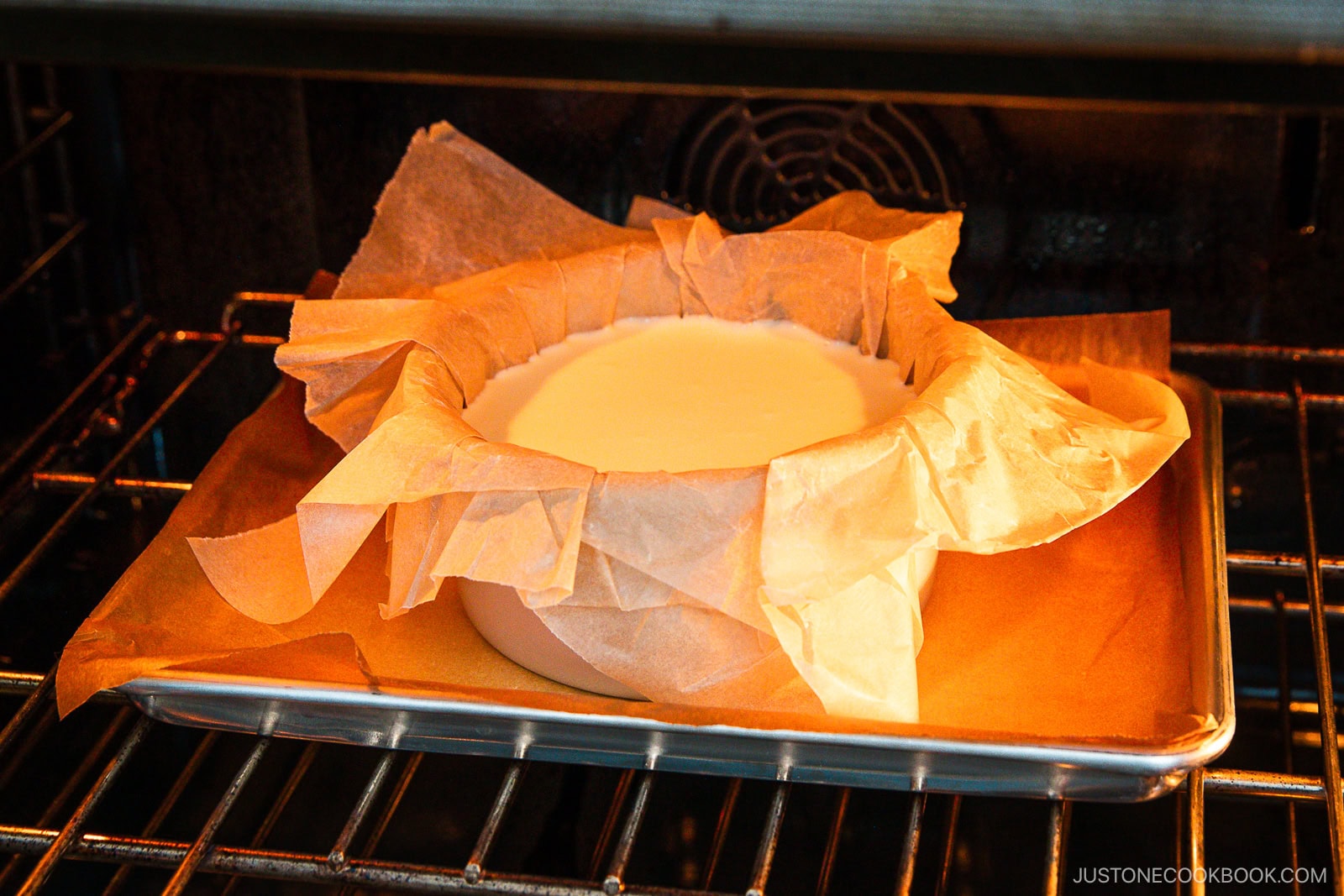 A round cake pan lined with parchment paper filled with batter is baking on a metal tray in an oven.