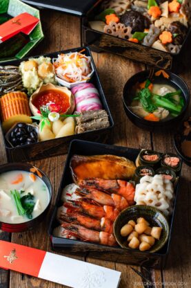 A colorful arrangement of traditional Japanese New Year&rsquo;s foods, including shrimp, fish, vegetables, and assorted delicacies in lacquered boxes, with bowls of soup on a wooden table.