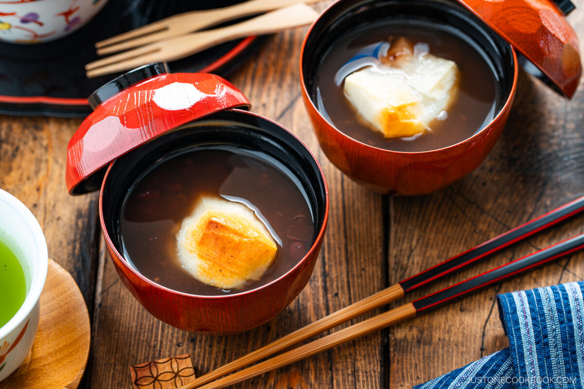 Two bowls of sweet red bean soup topped with grilled mochi sit on a wooden table, accompanied by a cup of green tea, chopsticks, and a blue-striped cloth.