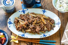 A platter of Beef and Gobo Stir Fry, topped with sesame seeds and served with chopsticks, a bowl of white rice, a cup, and small side dishes on a rustic wooden table.