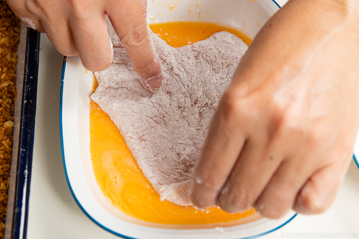 A person dips a flour-coated slice of meat into a dish of beaten eggs, preparing baked chicken katsu for breading. The hands and food are shown close up.