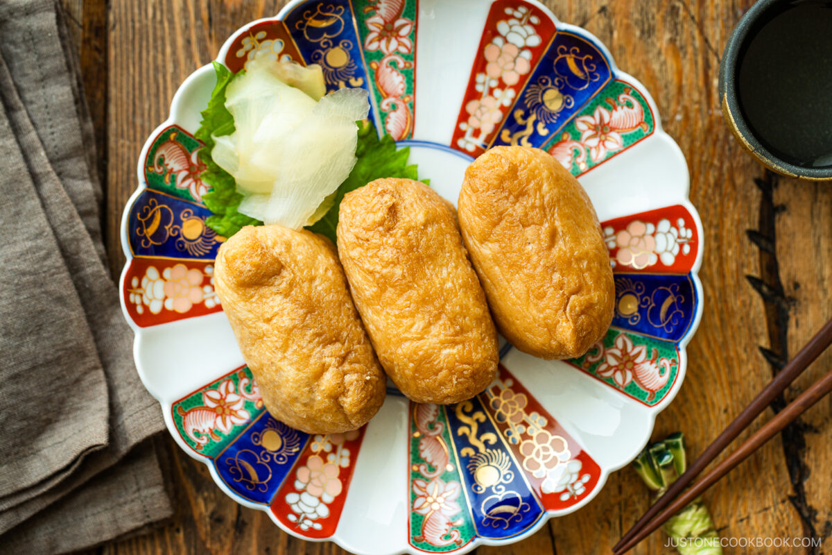 Three pieces of inari sushi (rice wrapped in fried tofu skin) are arranged on a colorful, decorative plate with pickled ginger and a lettuce leaf, set on a wooden table beside chopsticks and a brown napkin.