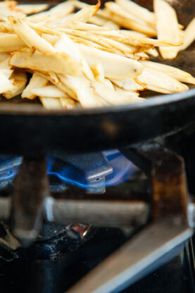 Sliced burdock root being saut&eacute;ed in a black skillet over a blue gas flame on a stovetop.
