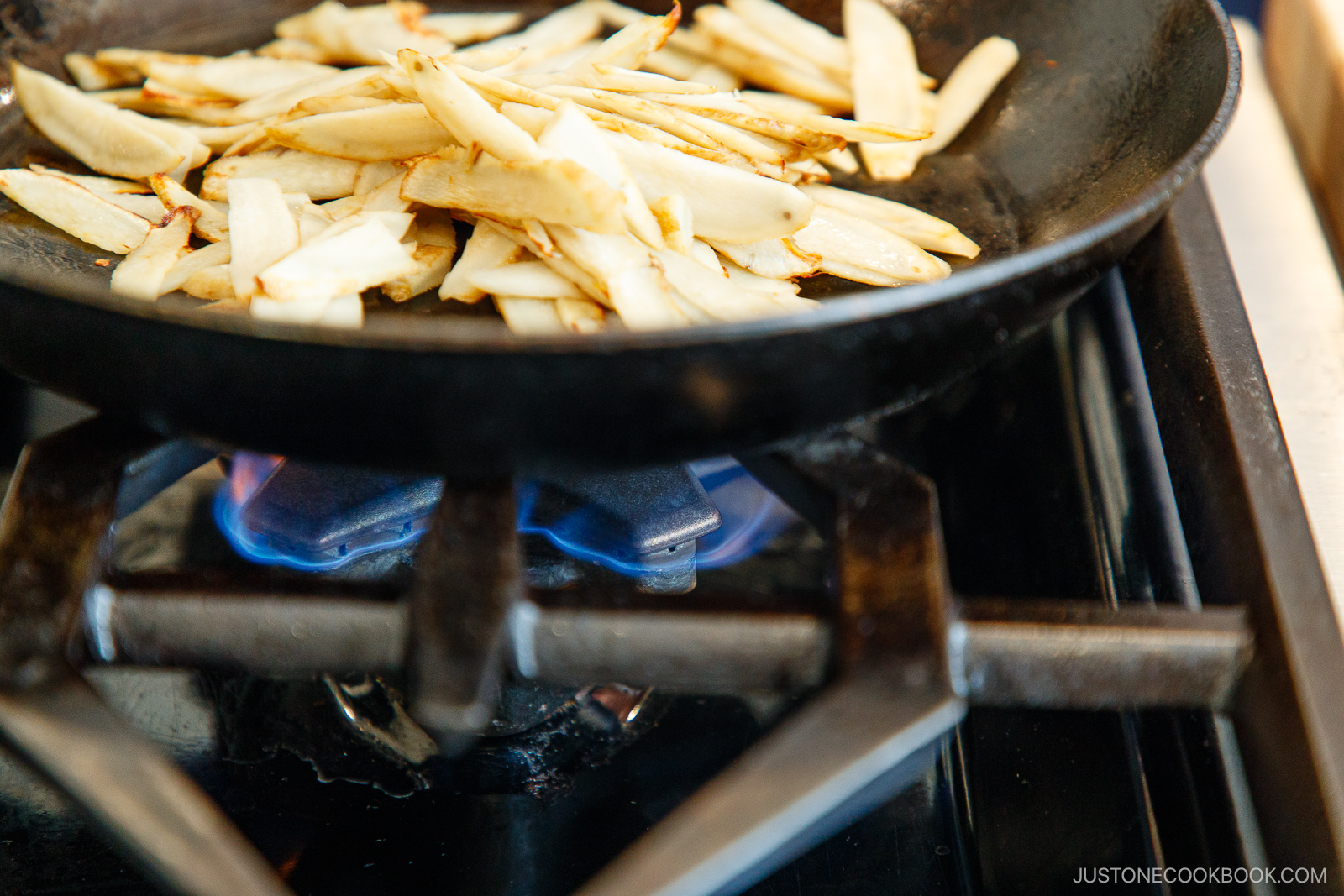 Sliced burdock root being saut&eacute;ed in a black skillet over a blue gas flame on a stovetop.