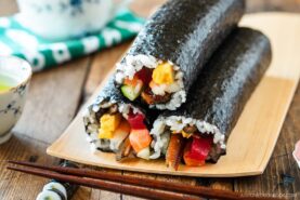 Two uncut sushi rolls&mdash;ehomaki (setsubun sushi roll)&mdash;filled with rice, vegetables, and seafood are wrapped in nori and arranged on a bamboo plate with chopsticks and a small teacup nearby.